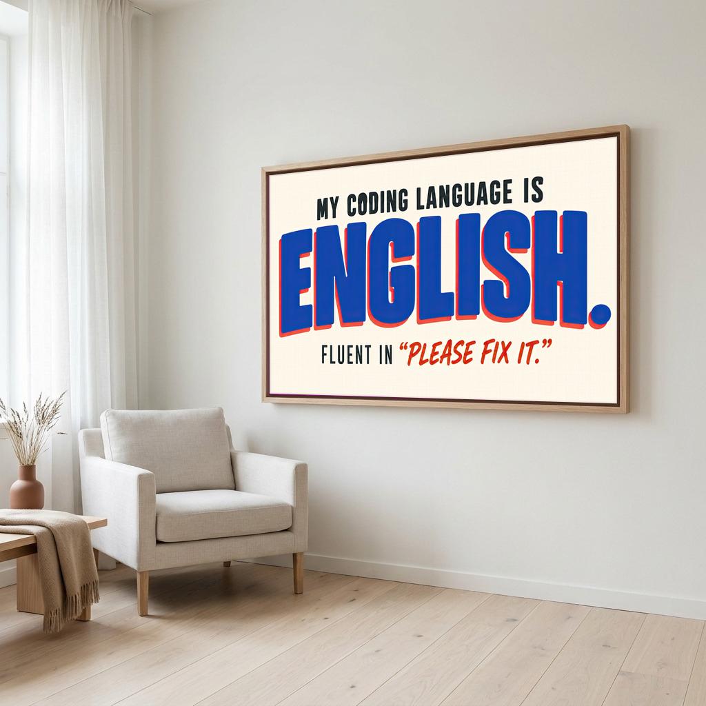 a large framed poster with the text "MY CODING LANGUAGE IS ENGLISH. FLUENT IN 'PLEASE FIT IT'". The poster is displayed on a wall in a room with a white chair and a wooden table.