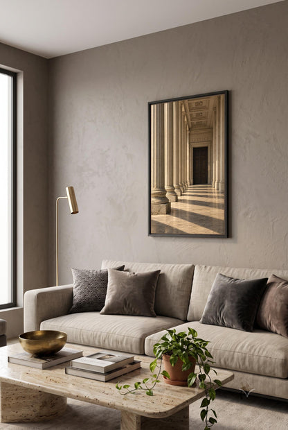 a modern living room with a large window, a gray sofa with throw pillows, a wooden coffee table, and a framed picture of a hallway with columns hanging on the wall.