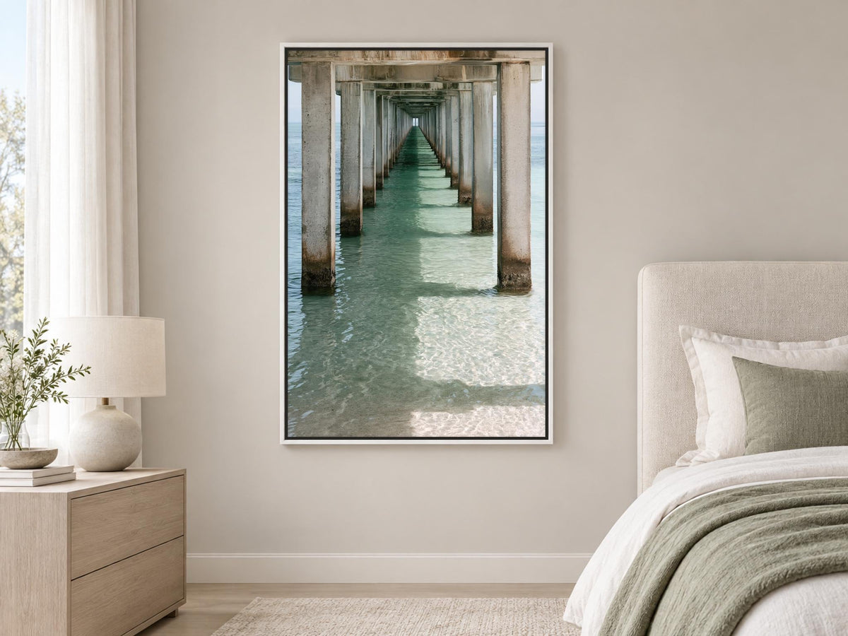 a serene bedroom scene with a large framed picture of a long pier or walkway extending into the water, creating a sense of depth and perspective. The room has a neutral color scheme with a white headboard and bedding, and a lamp on a side table.