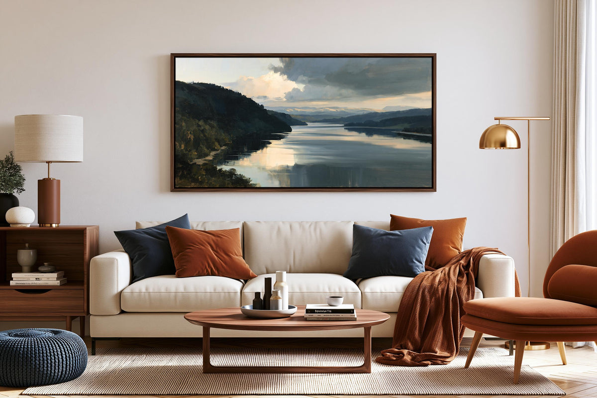 a cozy living room with a large painting of a serene landscape hanging on the wall above a white sofa adorned with throw pillows. The room is furnished with a wooden coffee table, a brown armchair, and a wooden side table with a lamp. The overall decor has a warm and inviting atmosphere, with the painting adding a touch of nature and tranquility to the space.
