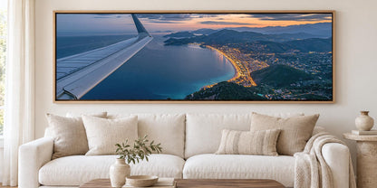 A large framed aerial photograph of a coastal city at sunset, with the wing of an airplane visible in the top left corner.