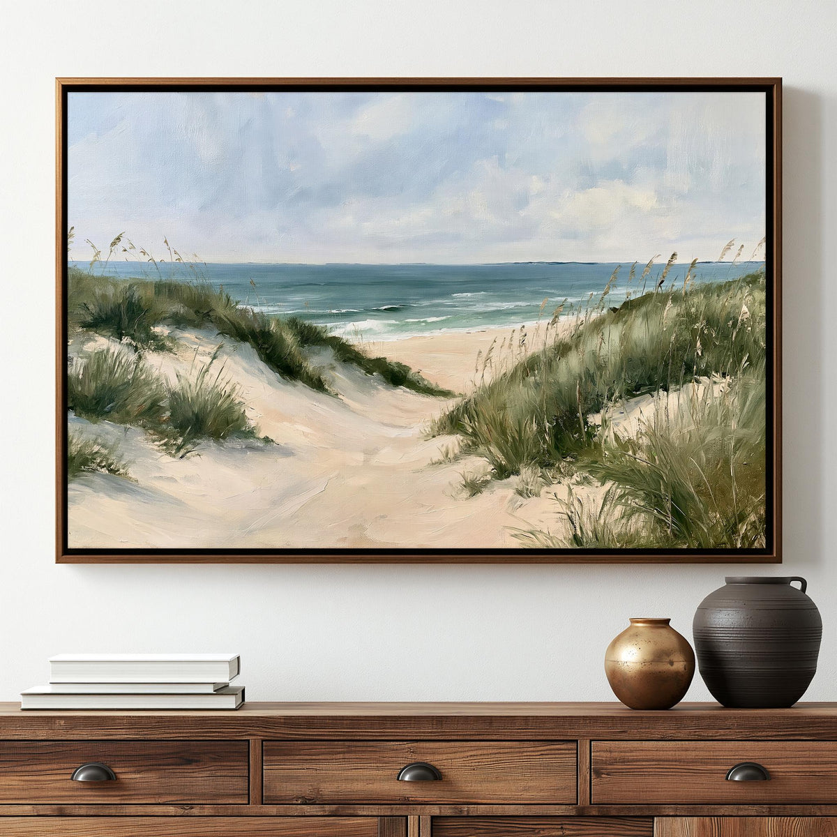 a large painting of a beach scene hanging on a wall above a wooden dresser. The painting depicts a sandy beach with dunes, grass, and the ocean in the background. The dresser has a stack of books and two decorative vases on it.