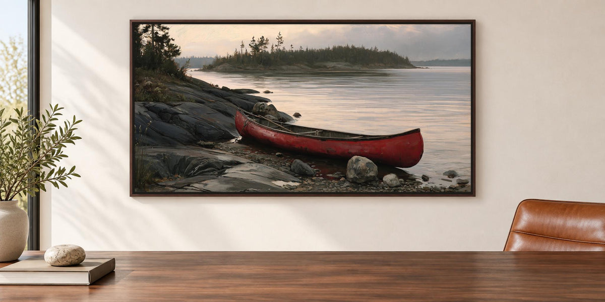 A framed painting of a red canoe resting on a rocky shore, with a calm lake and trees in the background.