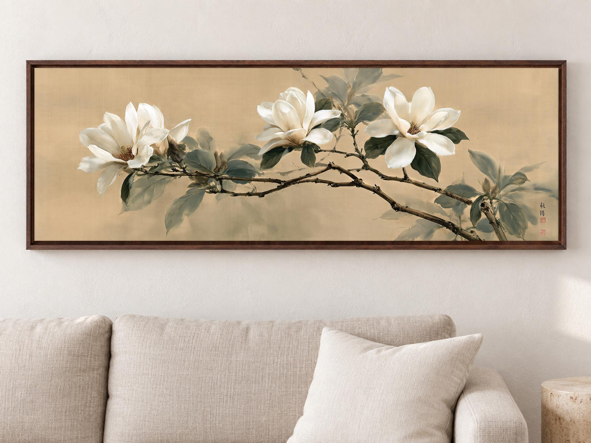 A framed painting of white flowers on a branch hangs on a wall above a beige couch in a living room.
