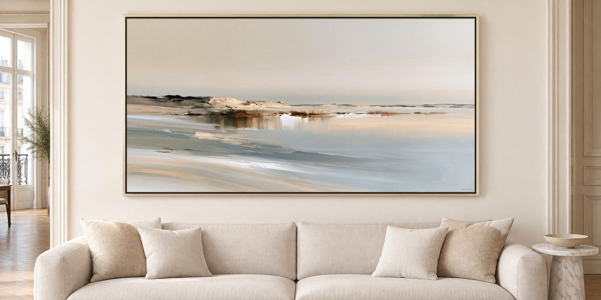 a large, framed painting of a serene landscape hanging on a wall above a beige sofa with two pillows.