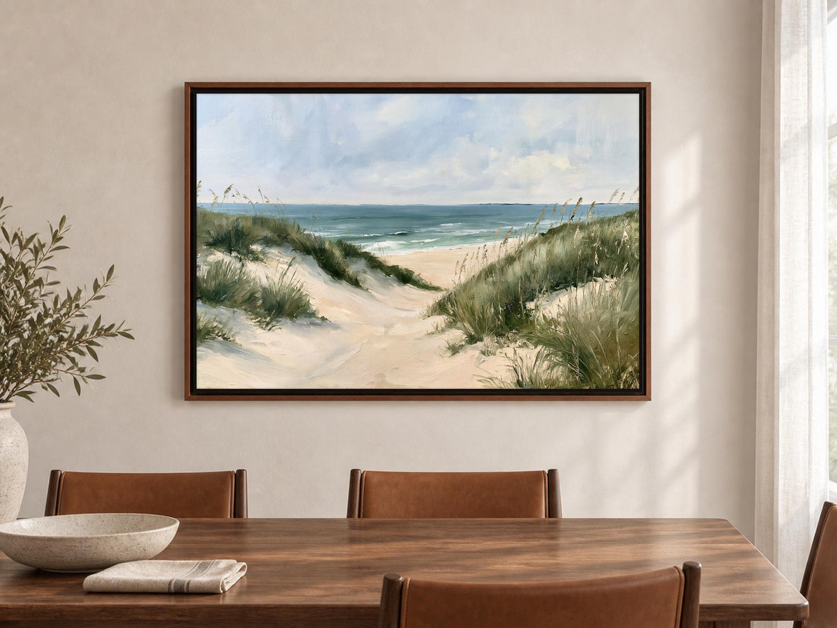a large framed painting of a beach scene with sand dunes and grassy areas, hanging on a wall above a wooden dining table with four chairs.
