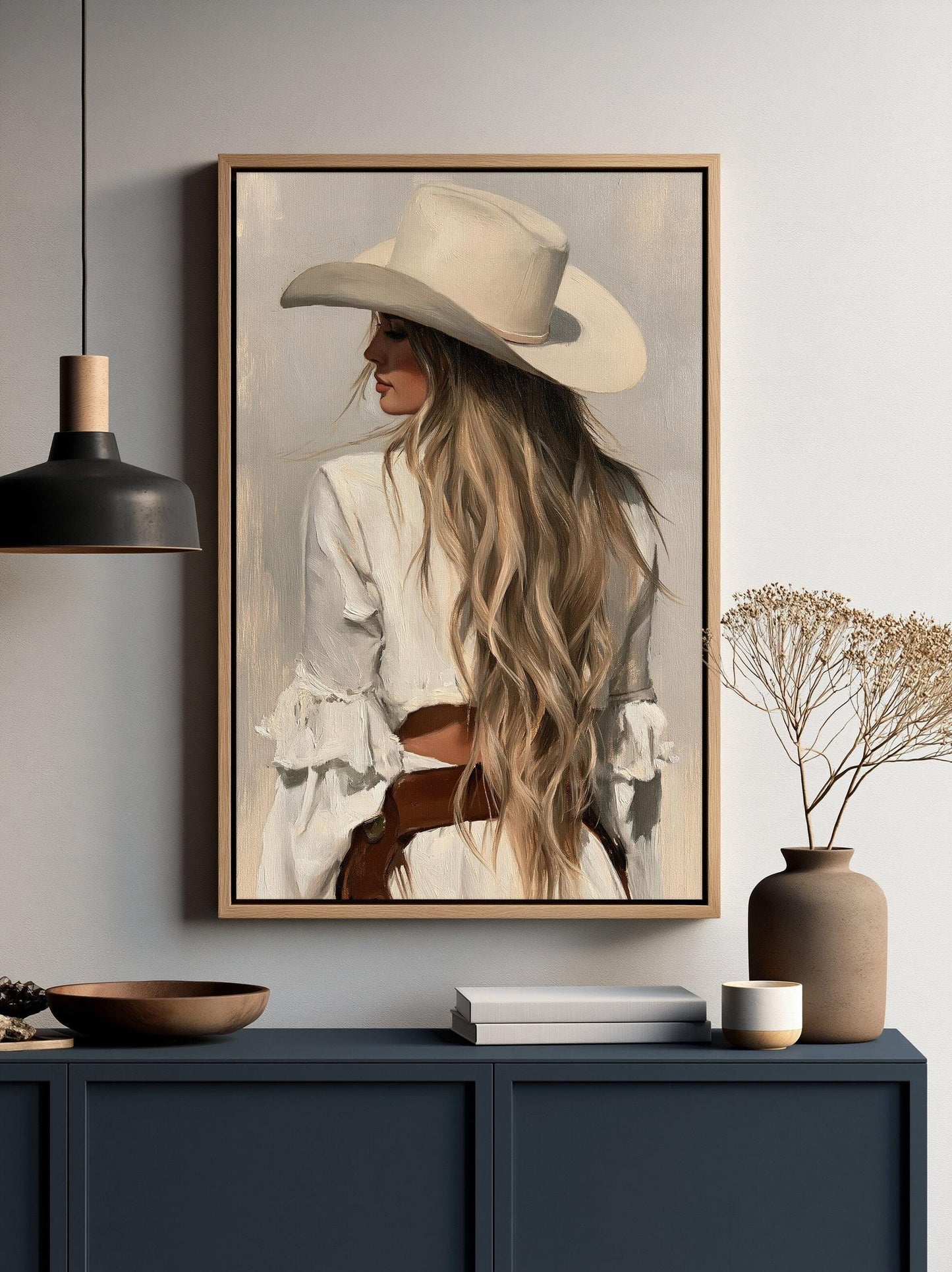 a framed painting of a woman with long blonde hair wearing a white hat, displayed on a wall above a dark blue cabinet.