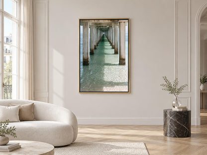 a serene indoor setting with a large window overlooking a long pier extending into the water. The pier is lined with pillars and features a view of the ocean. The room is decorated with a white sofa, a round coffee table, and a marble side table with a plant. The overall atmosphere is calm and peaceful, with the natural elements of the pier and the ocean providing a sense of tranquility.