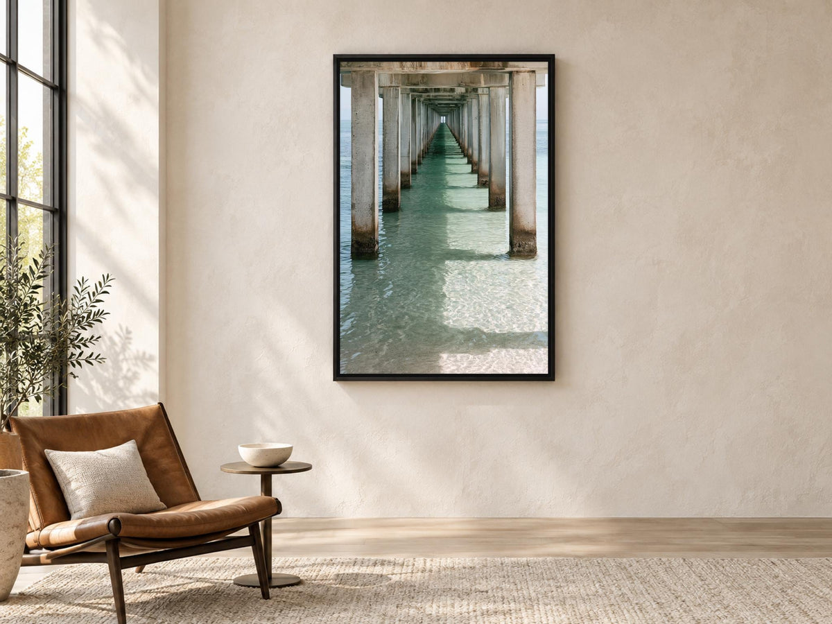a serene indoor setting with a large framed photograph of a long pier or walkway extending into the distance, creating a sense of depth and perspective. The photograph is hung on a light-colored wall, and the room is furnished with a brown leather chair, a small side table, and a potted plant, creating a cozy and inviting atmosphere.