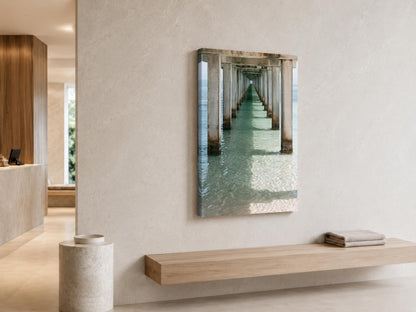 a minimalist interior design with a large wall-mounted photograph of a long pier or walkway extending into the distance. The photograph is framed in a rectangular canvas and hangs on a beige wall. In front of the wall, there is a wooden bench and a small white pedestal. The overall color scheme of the room is neutral, with the beige wall and wooden elements providing a calming and serene atmosphere.