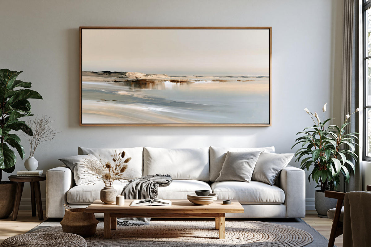 a serene living room with a large painting of a beach scene hanging on the wall above a white sofa. The sofa is adorned with a throw blanket and a vase of dried flowers. The room also features a wooden coffee table, a potted plant, and a large window that allows natural light to fill the space.