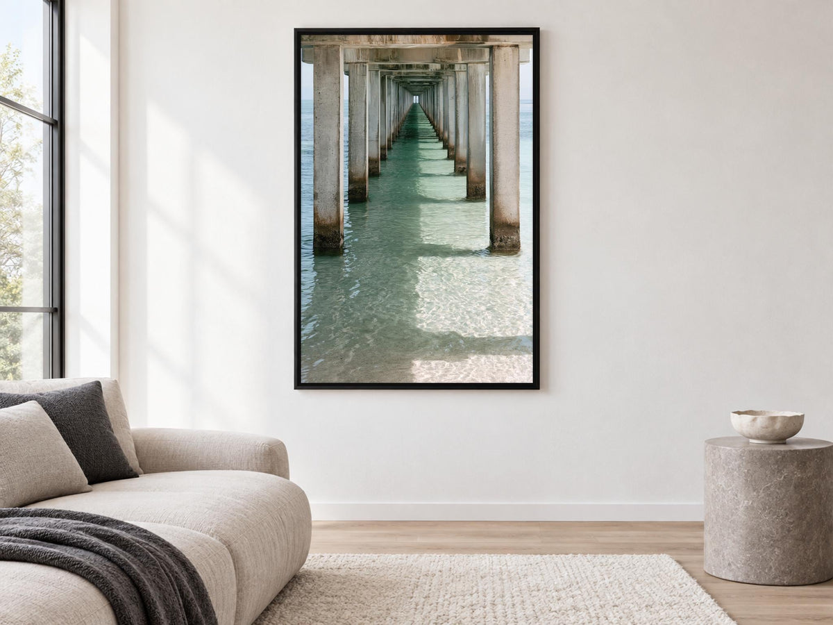 a large framed picture of a long pier with a view of the water, hanging on a white wall in a living room.