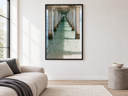 a large framed picture of a long pier with a view of the water, hanging on a white wall in a living room.