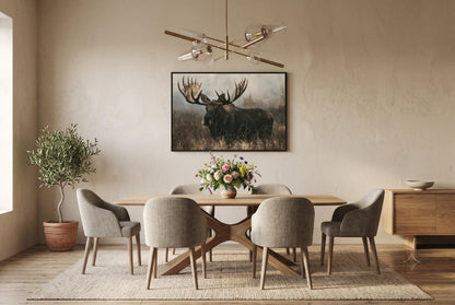 a dining room with a wooden table and six upholstered chairs. Above the table, there is a framed picture of a moose. The room has a neutral color scheme with beige walls and a wooden floor. A chandelier is hanging from the ceiling, and there is a potted plant and a vase on the table.