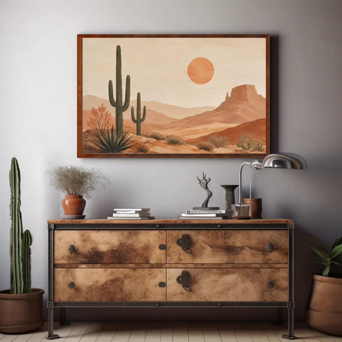 a framed desert landscape painting hanging on a wall above a wooden dresser with drawers. The painting depicts a desert scene with cacti, mountains, and a large orange sun in the sky. The dresser has various decorative items on it, including a potted plant, a vase, and a lamp.