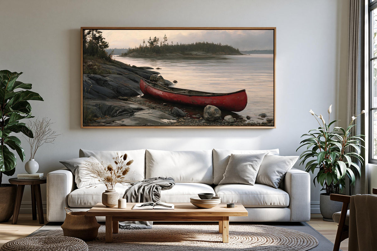 a large, framed painting of a red canoe resting on a rocky shore, with a serene lake and trees in the background. The painting is hung on a wall above a white sofa in a cozy living room setting.