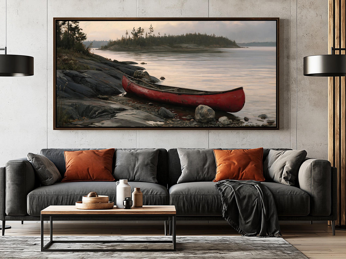 a large, framed painting of a red canoe resting on a rocky shore, with a calm lake and trees in the background. The painting is hung on a wall above a gray sofa with orange throw pillows.