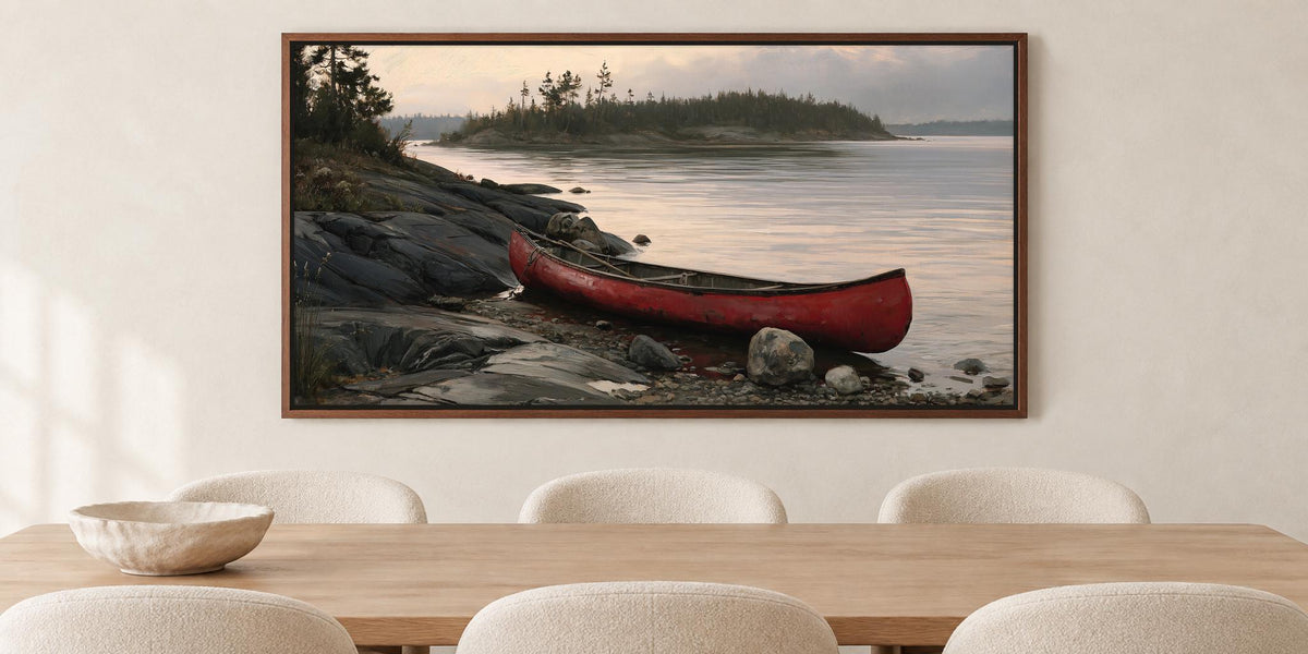 A framed painting of a red canoe resting on a rocky shore, with a calm lake and trees in the background, hangs on a wall above a wooden dining table with white chairs.