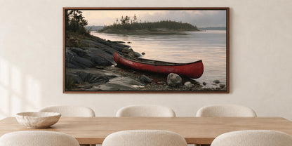 A framed painting of a red canoe resting on a rocky shore, with a calm lake and trees in the background, hangs on a wall above a wooden dining table with white chairs.
