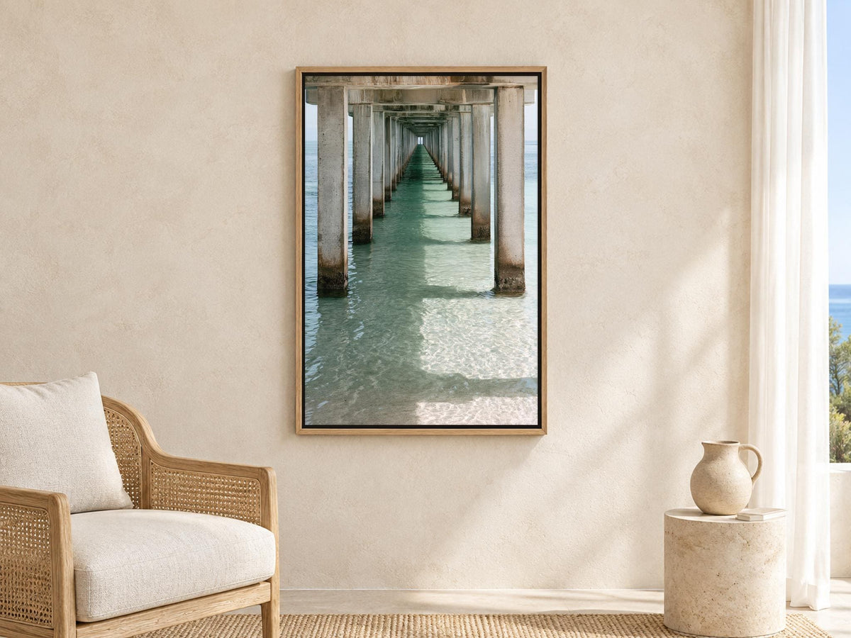 a serene indoor setting with a large framed picture of a long pier or walkway extending into the water, creating a sense of depth and perspective. The pier is supported by numerous columns, adding architectural interest to the scene. The photograph is hung on a beige wall, which complements the overall color scheme of the room. In the foreground, there is a wicker chair and a small vase, adding to the cozy and inviting atmosphere of the space.