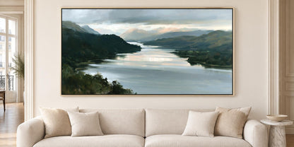 a large painting of a serene landscape with a river and mountains in the background, hanging on a wall above a beige sofa with pillows.