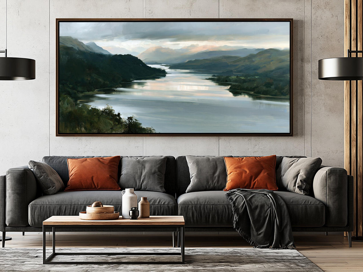 a large painting of a serene landscape with a river and mountains, hanging on a wall above a gray sofa with orange throw pillows. The painting is framed in a dark wood frame, and the sofa is positioned in front of it, creating a cozy and inviting atmosphere in the living room.