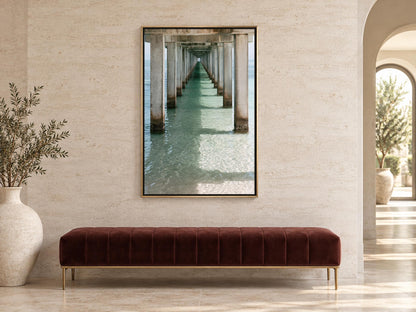 a long, straight pier extending into the water, with a series of pillars supporting the structure. The pier is surrounded by a serene, natural environment, with the water appearing calm and inviting. The photograph is framed and hung on a wall, creating a focal point in the room.