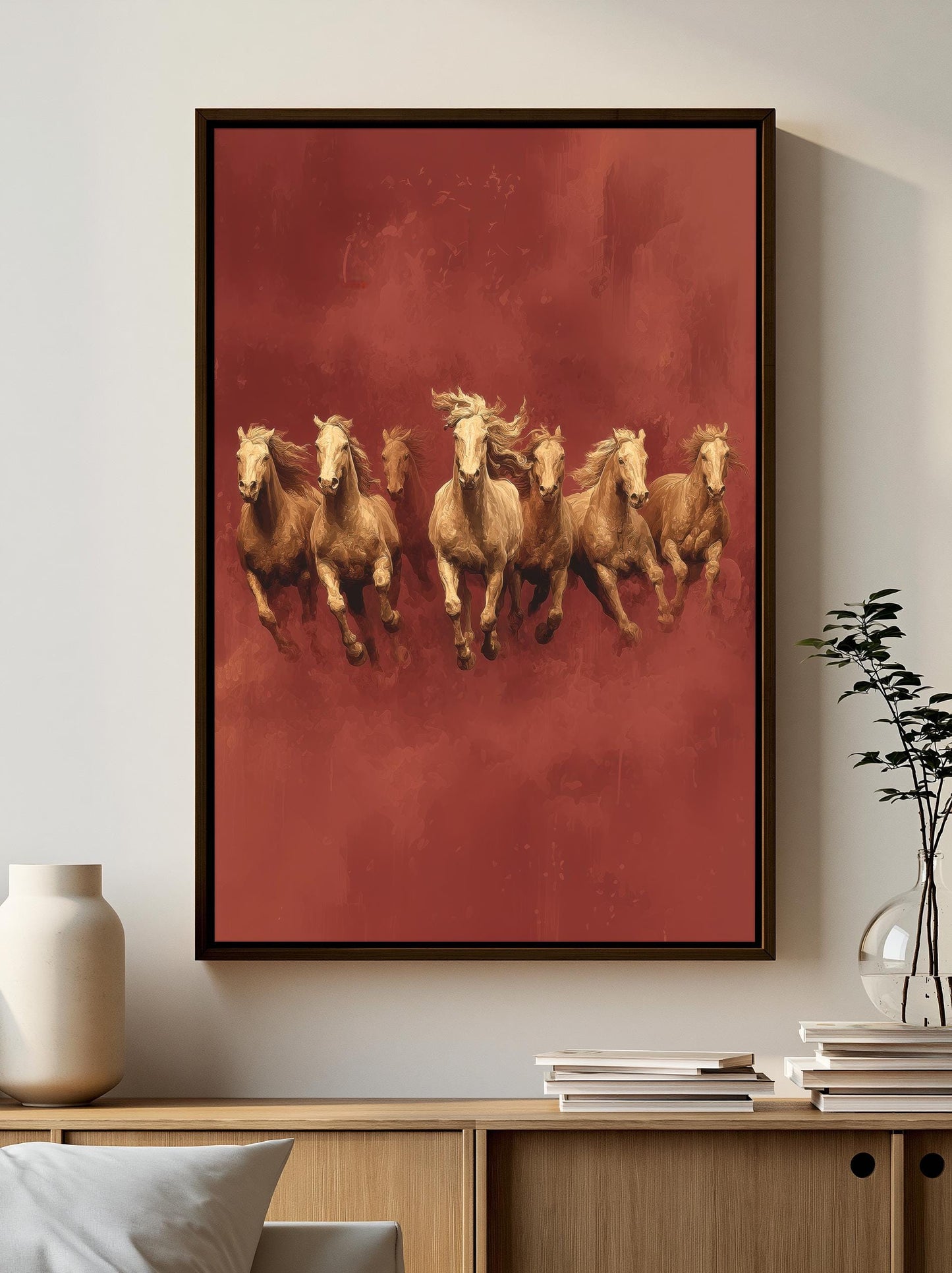A framed painting of six galloping horses in a red background hangs on a wall, with a wooden cabinet below it and a stack of books on top.