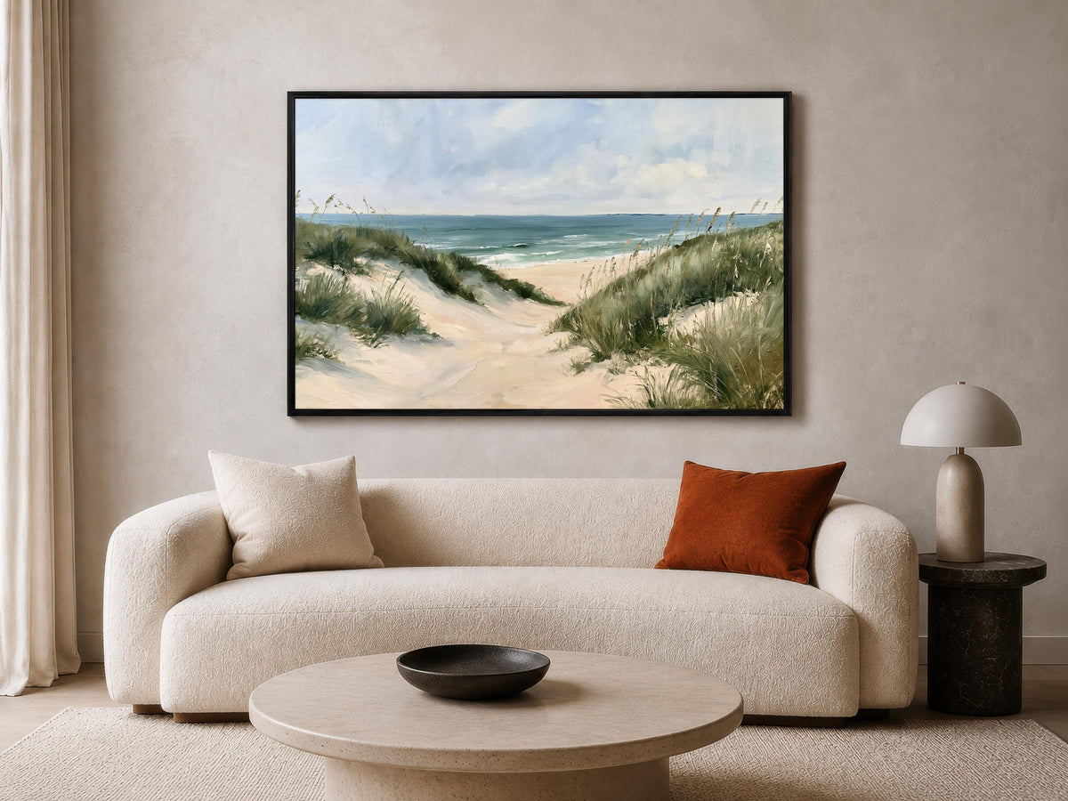 a living room with a large painting of a beach scene hanging on the wall above a white sofa. The painting depicts a sandy beach with dunes and grass, and the ocean in the background. The room has a modern and minimalist design, with a round coffee table in the center and a lamp on a side table.
